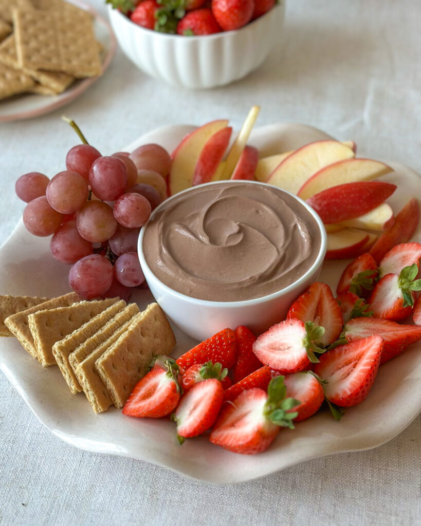 High-protein chocolate dip surrounded by fruit and graham crackers with a bowl of strawberries in the background