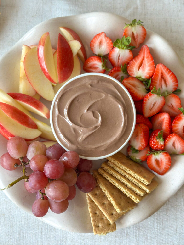 High-protein chocolate dip surrounded by strawberries, apple slices, red grapes, and graham crackers on a platter