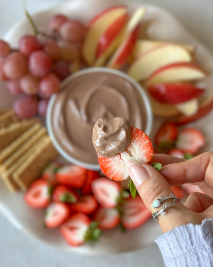 Hand holding a strawberry topped with high-protein chocolate dip with the fruit platter blurred in the background