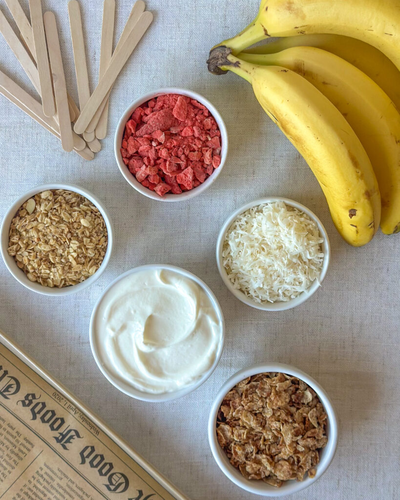 Ingredients for frozen Greek yogurt banana pops including bananas, popsicle sticks, Greek yogurt, crushed cereal, granola, freeze-dried strawberries, and coconut