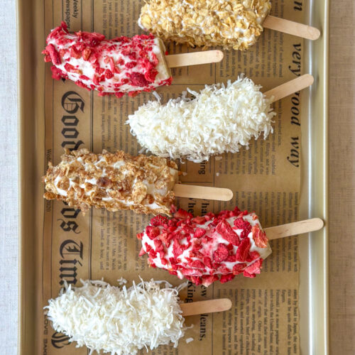Overhead view of frozen Greek yogurt banana pops covered with colorful toppings on a parchment-lined golden