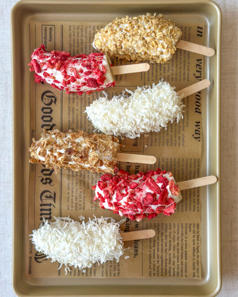 Overhead view of frozen Greek yogurt banana pops covered with colorful toppings on a parchment-lined golden
