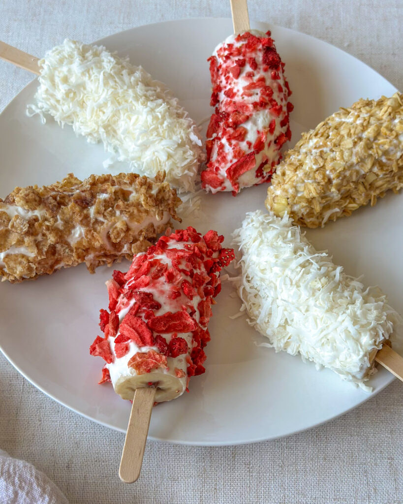 Close-up of colorful frozen banana pops coated in Greek yogurt and toppings arranged on a white plate