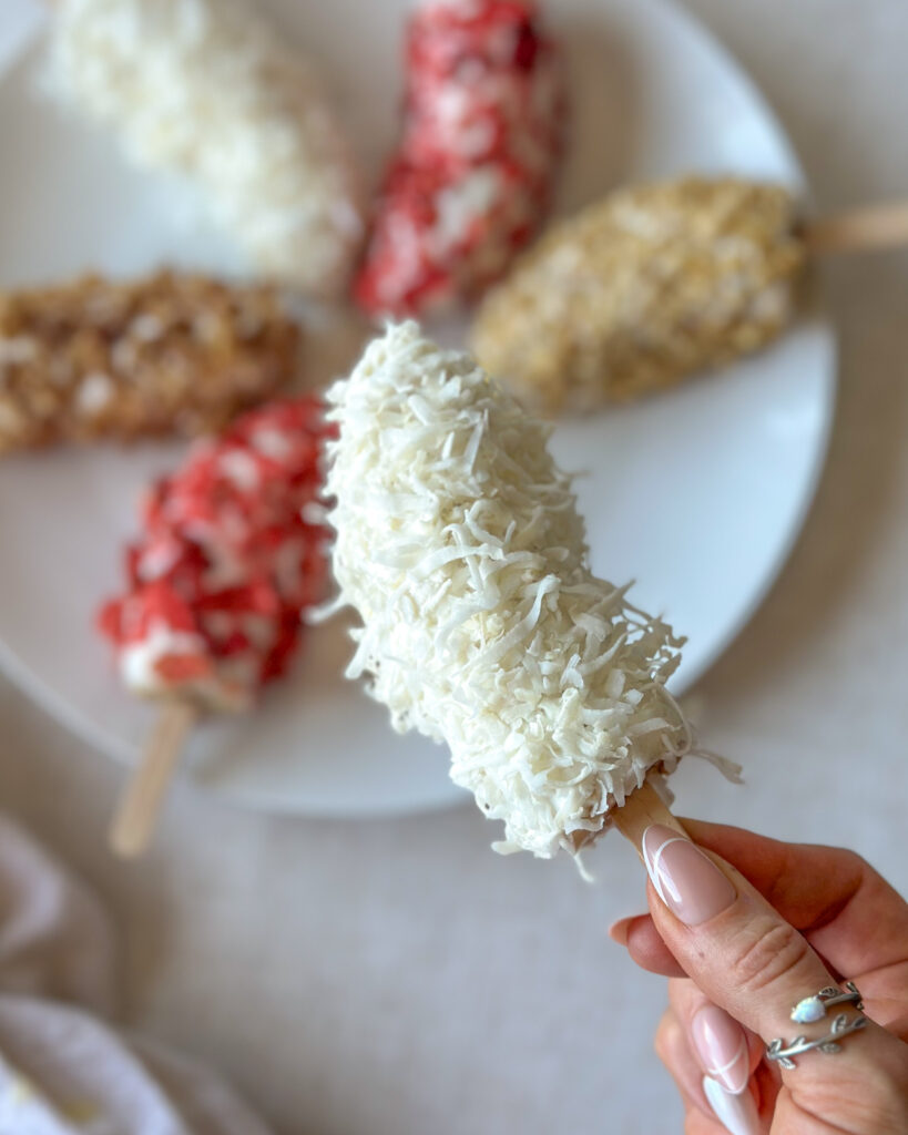 Hand holding an intact frozen Greek yogurt banana pop with more colorful banana pops arranged on a white plate in the background