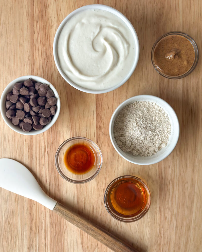 Flat lay of ingredients for high-protein cookie dough dip including oat flour, Greek yogurt, chocolate chips, maple syrup, and vanilla