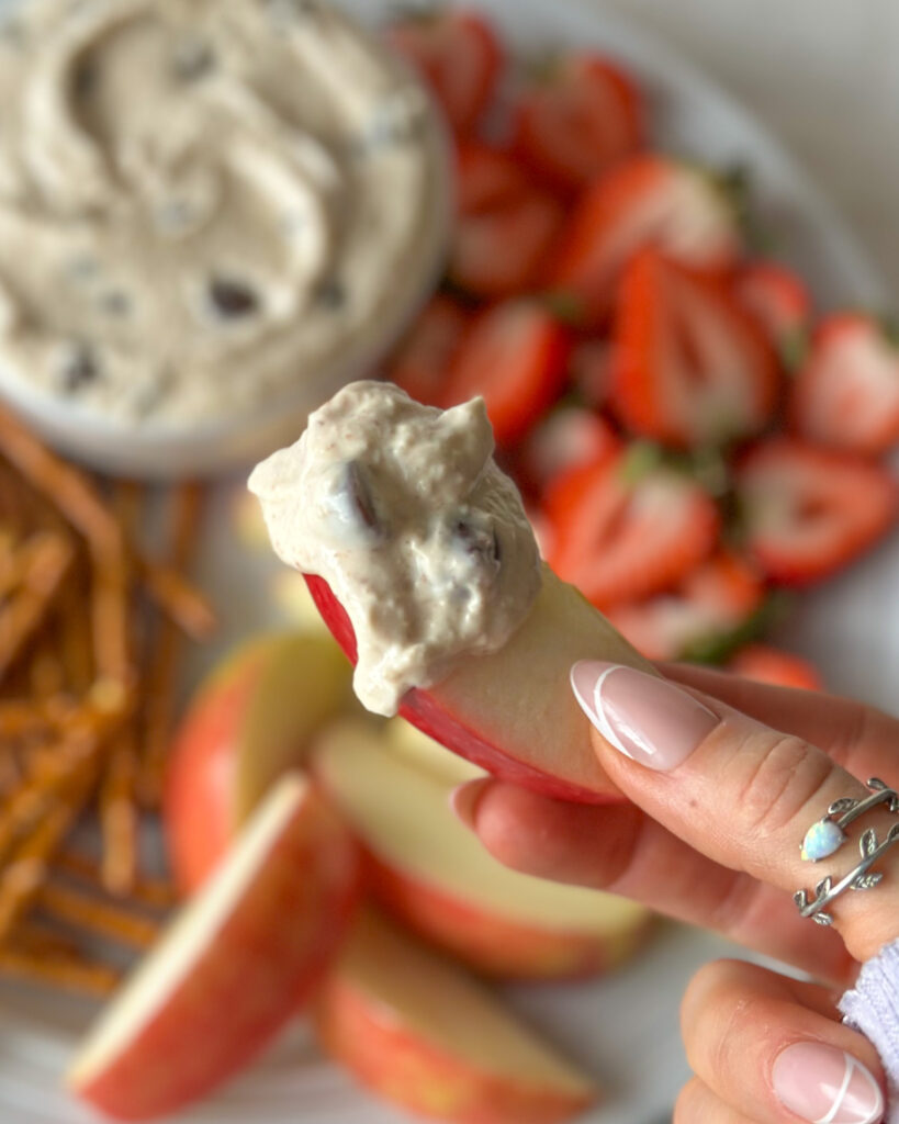 Hand holding an apple slice topped with a dripping dollop of high-protein cookie dough dip with the finished platter blurred in the background