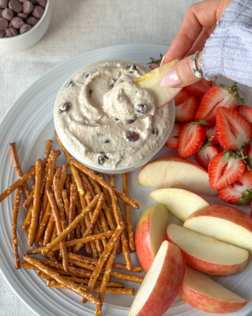 Finished high-protein cookie dough dip platter with chocolate chips in the background and an apple slice being dipped into the dip