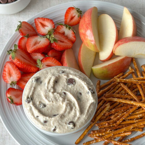 Overhead close-up of swirled high-protein cookie dough dip surrounded by strawberries, apple slices, and pretzels with chocolate chips in the background