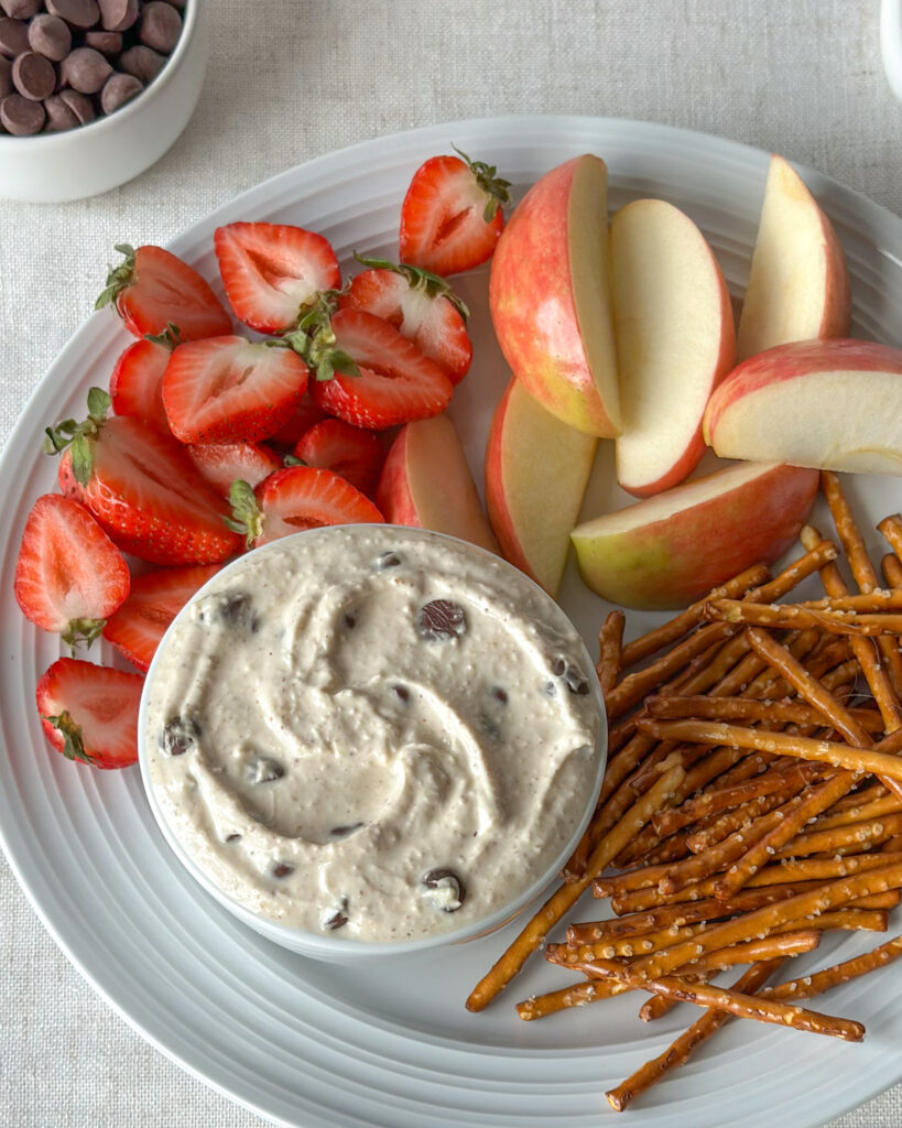 Overhead close-up of swirled high-protein cookie dough dip surrounded by strawberries, apple slices, and pretzels with chocolate chips in the background