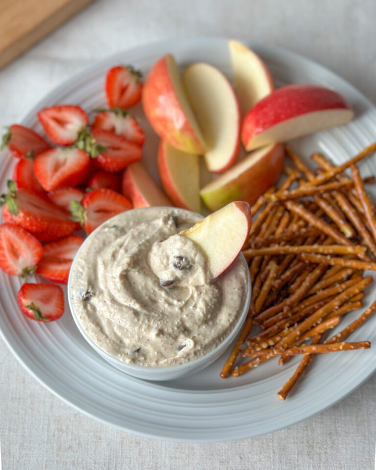 High-protein cookie dough dip surrounded by pretzels, apple slices, and strawberries with an apple slice propped in the center