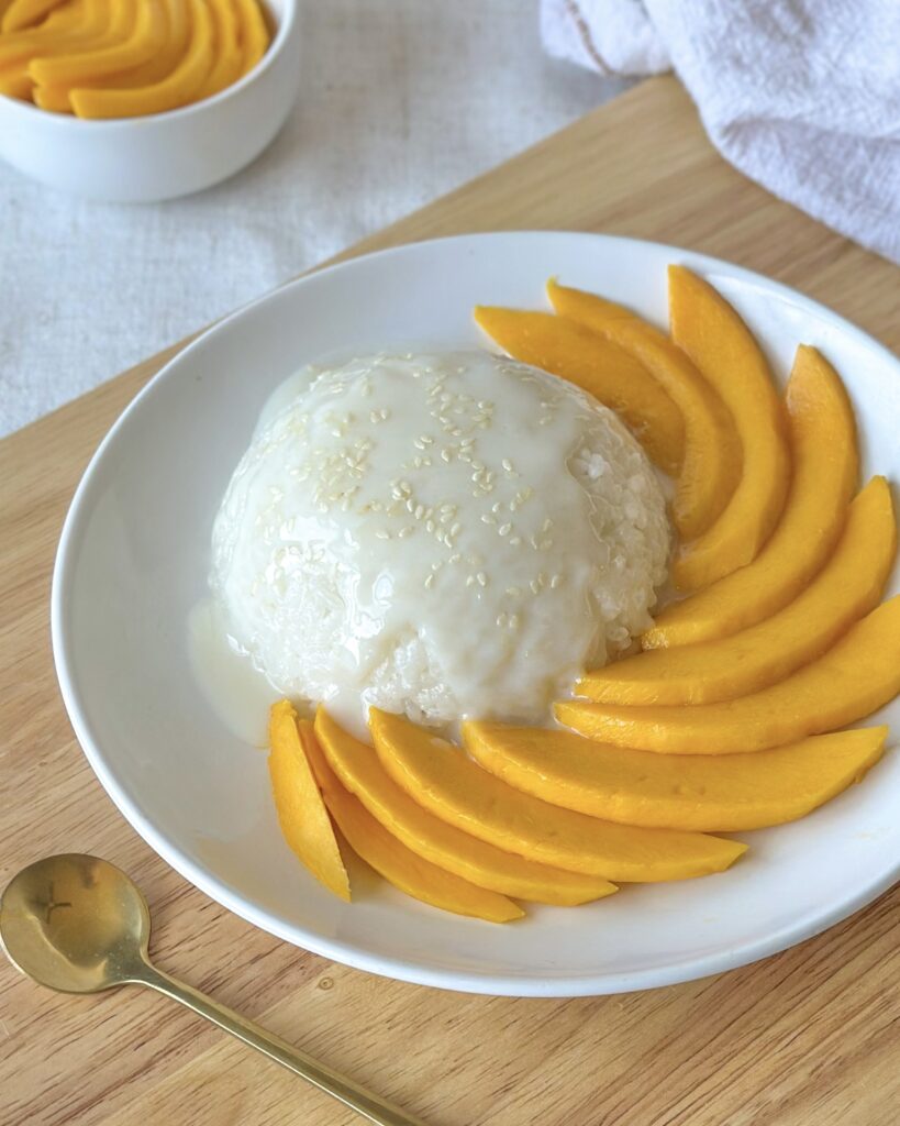 mango-sticky-rice-with-mango-slices.jpeg Mango sticky rice served with fresh mango at the front and a dome of sticky rice beside a spoon ready for serving.