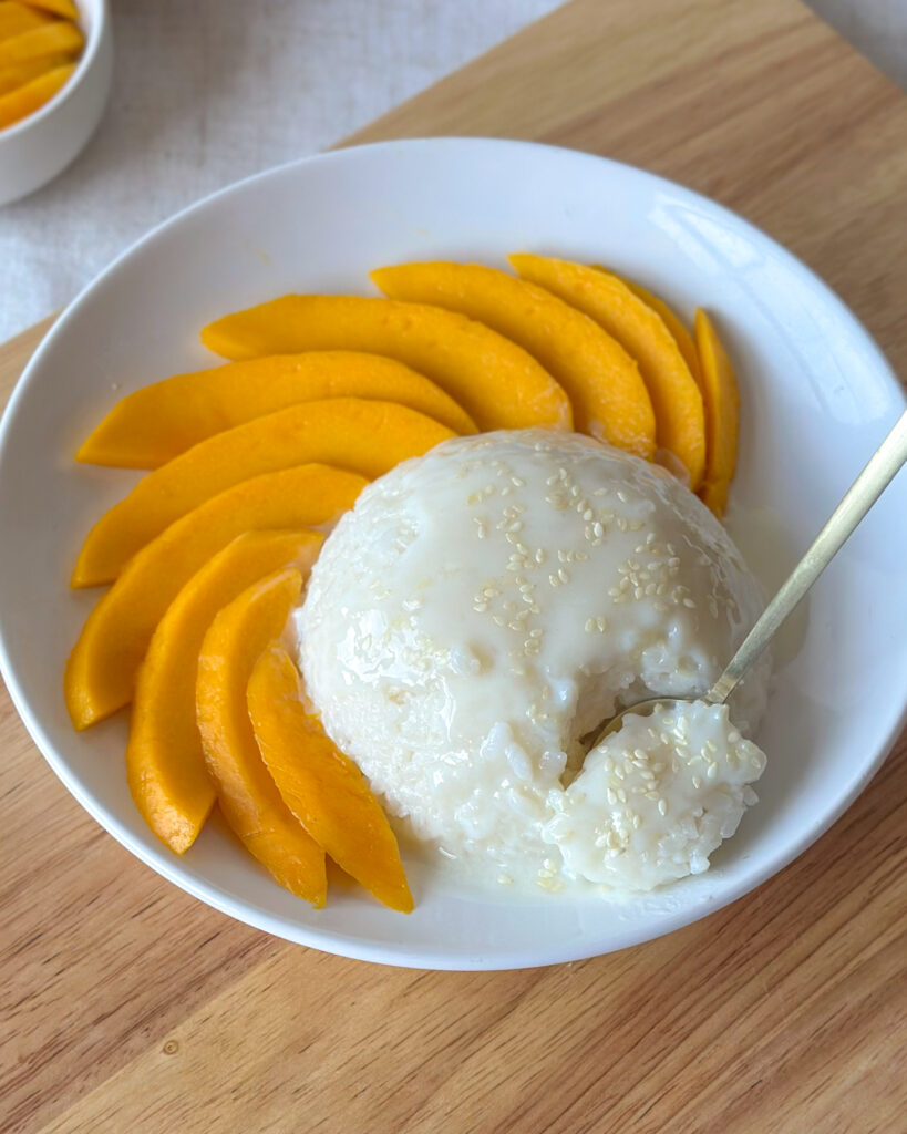 Spoon dipping into a spherical mound of mango sticky rice in a white shallow bowl, with fresh mango in the background on a wooden chopping board.