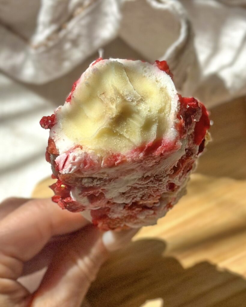 hand holding frozen banana pop with strawberry coating with the center revealed