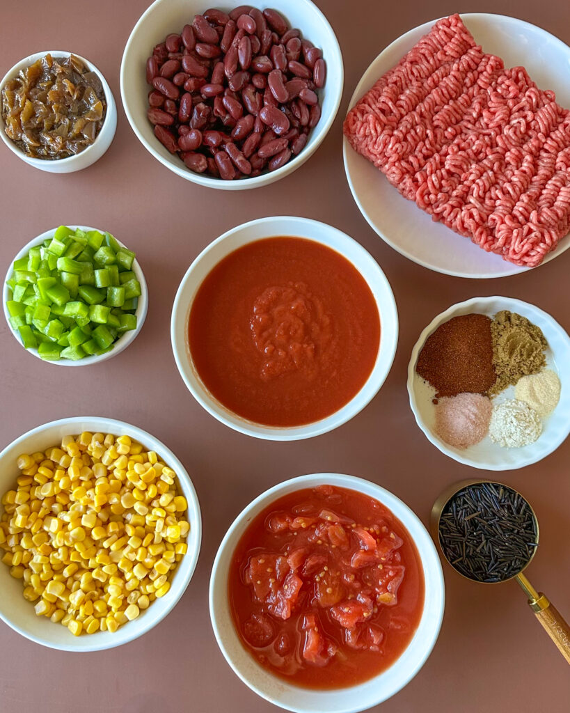 Flat lay of ingredients for wild rice chili including ground beef, wild rice, caramelized onions, hearts of palm, beans, corn, bell peppers, tomatoes, and spices