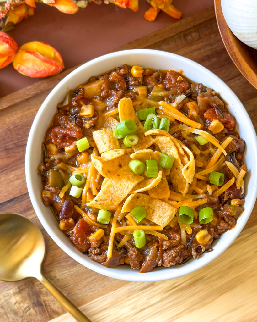 Overhead view of a bowl of homemade chili topped with cheese, corn chips, and green onion with garlic and flowers in the background