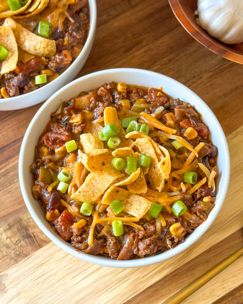 Overhead view of hearty homemade chili topped with melted cheese, corn chips, and green onion on a wooden board