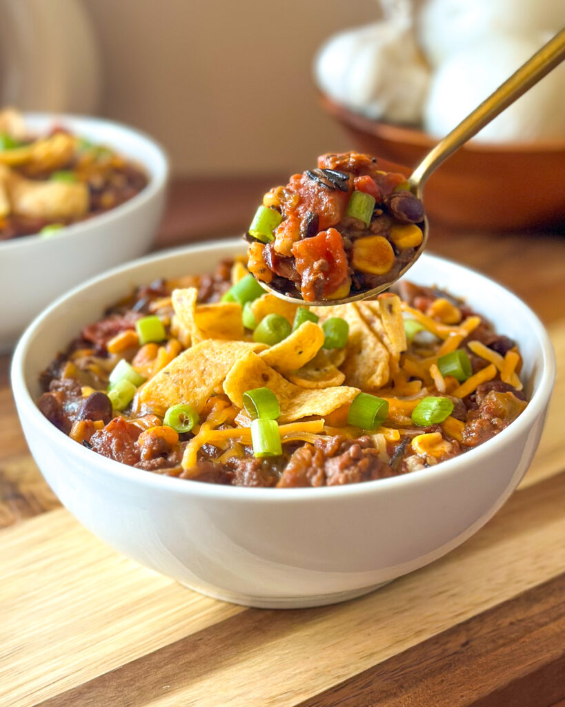 Spoon lifting a generous scoop of hearty wild rice chili over a bowl with another serving and garlic in the background