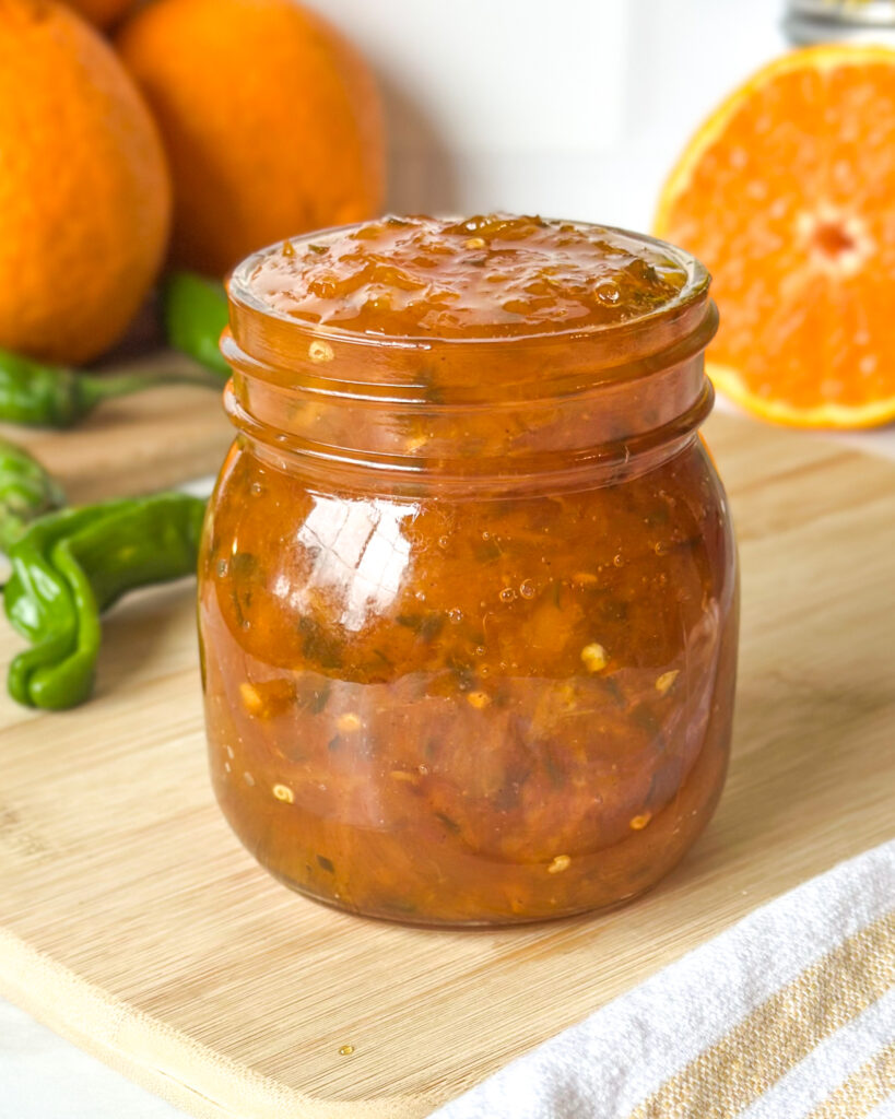 Front view of spicy citrus pepper jelly in an open glass jar showing the glossy texture with citrus and peppers in the background