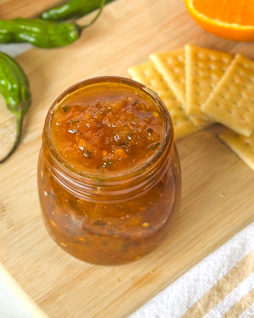 Overhead view of spicy citrus pepper jelly in an open glass jar showing its soft jammy texture with crackers and peppers in the background