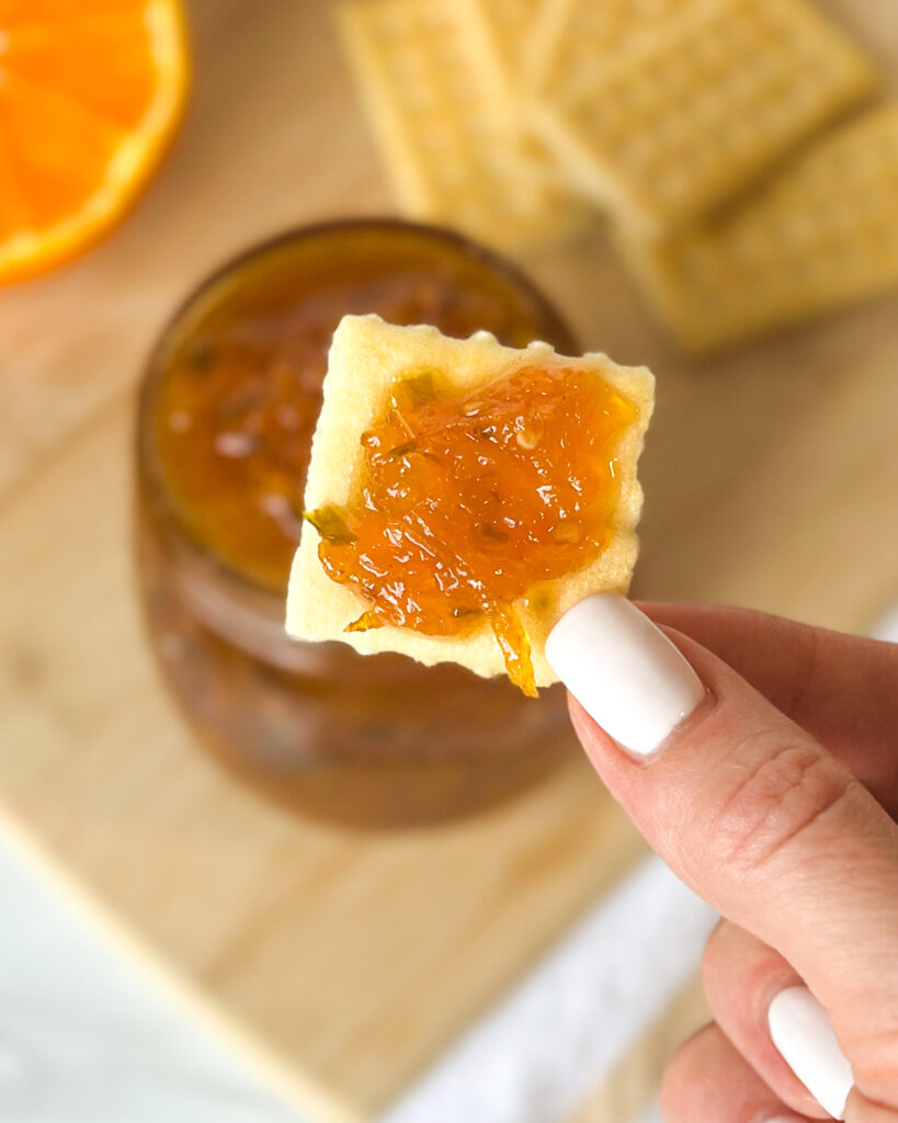 Fingers holding a cracker topped with spicy citrus pepper jelly with the jar, crackers and sliced Sumo citrus blurred in the background
