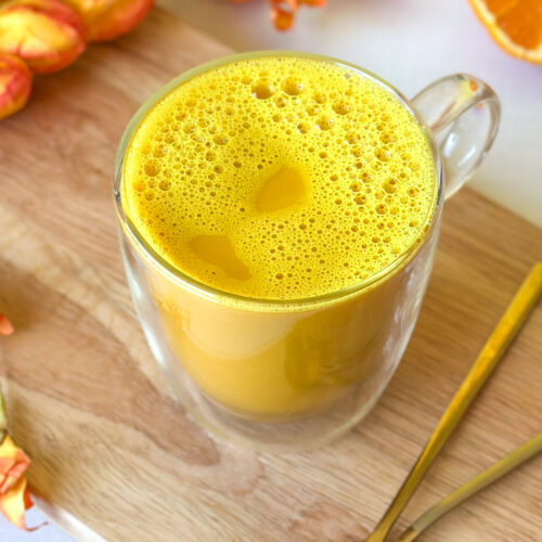 Golden milk turmeric latte with a frothy top in a clear glass mug surrounded by golden spoons, sliced Sumo citrus and orange and yellow flowers