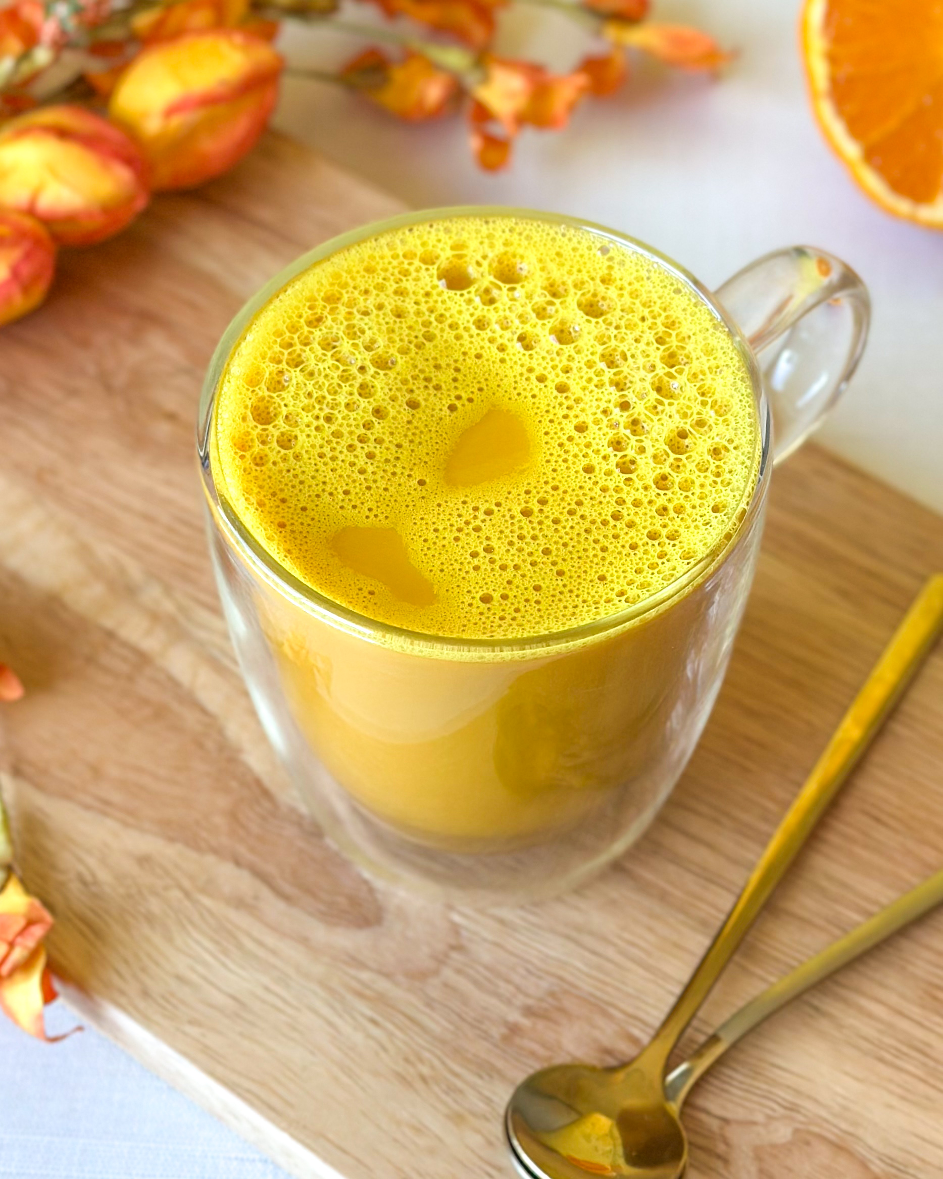 Golden milk turmeric latte with a frothy top in a clear glass mug surrounded by golden spoons, sliced Sumo citrus and orange and yellow flowers