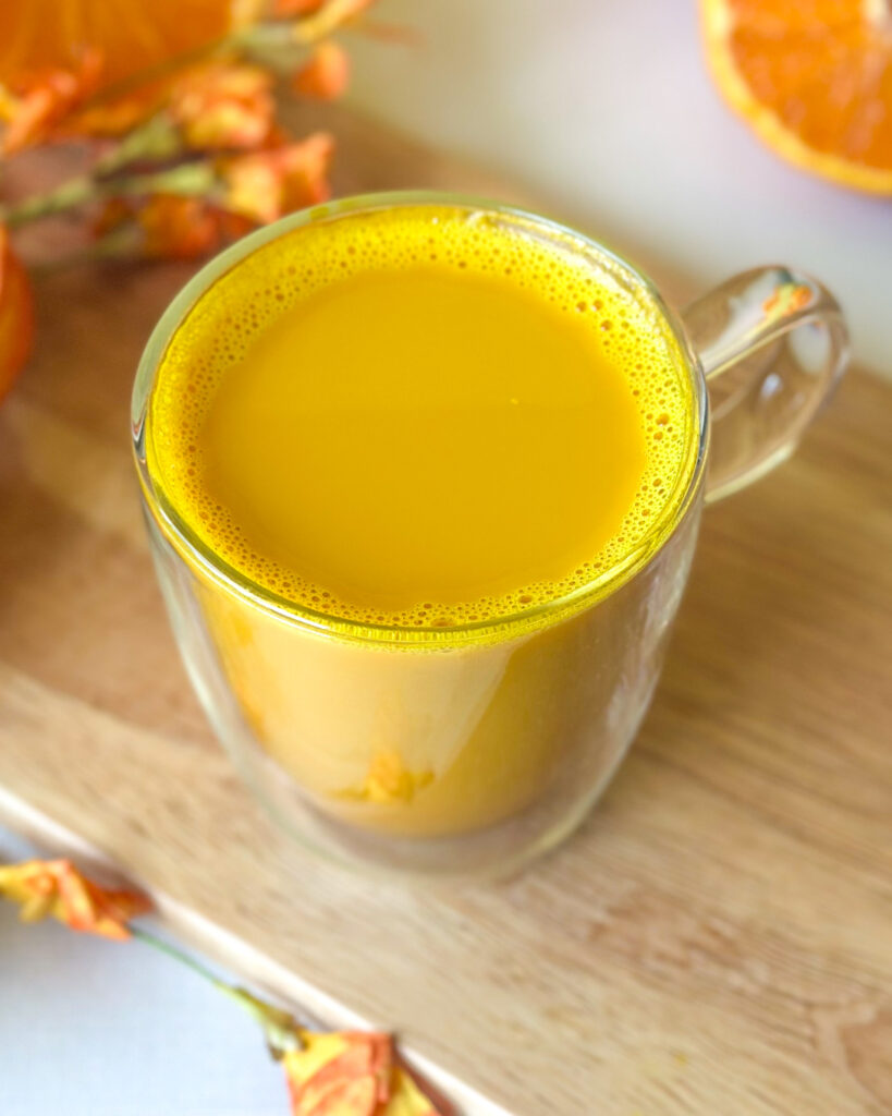 Golden milk turmeric latte in a clear glass mug with a frothy rim on a wooden board surrounded by golden flowers