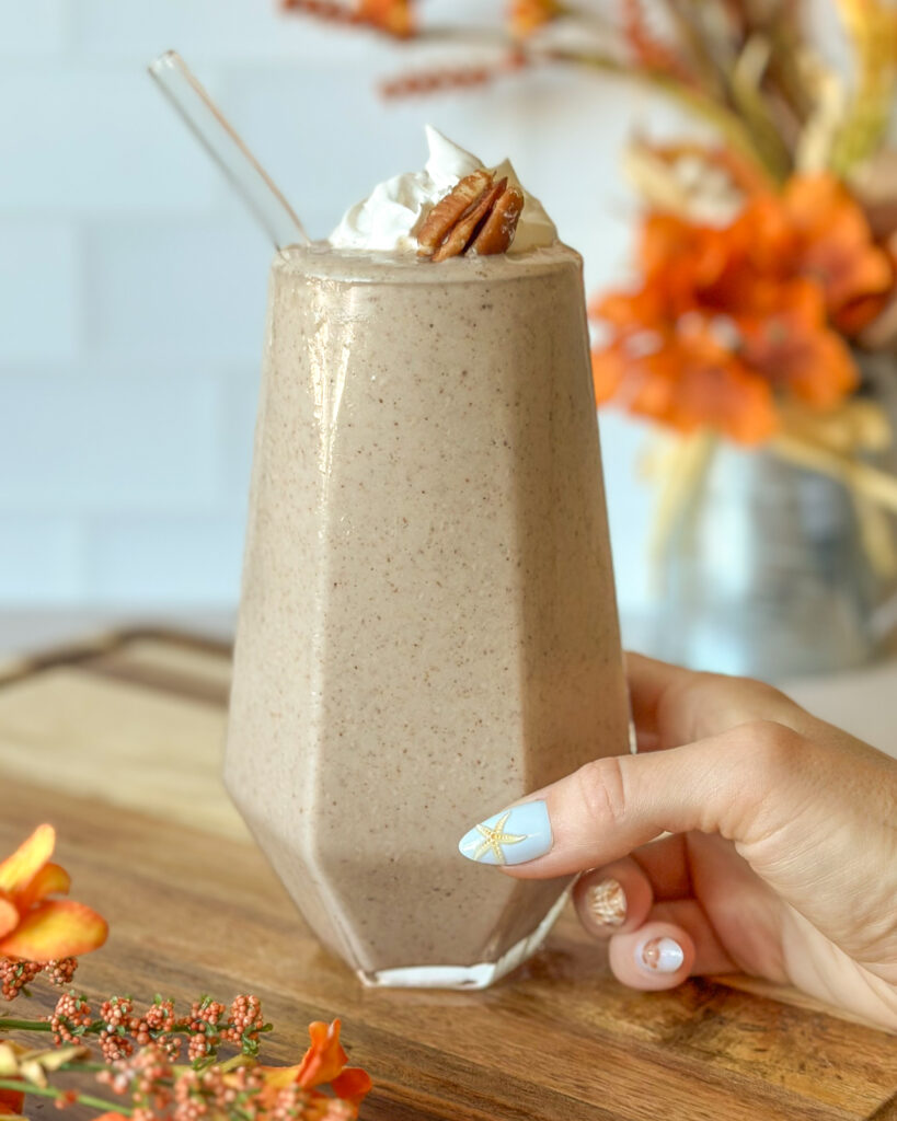 Hand holding a pecan pie smoothie in a tall geometric glass topped with whipped cream and a pecan, with fall flowers in the background