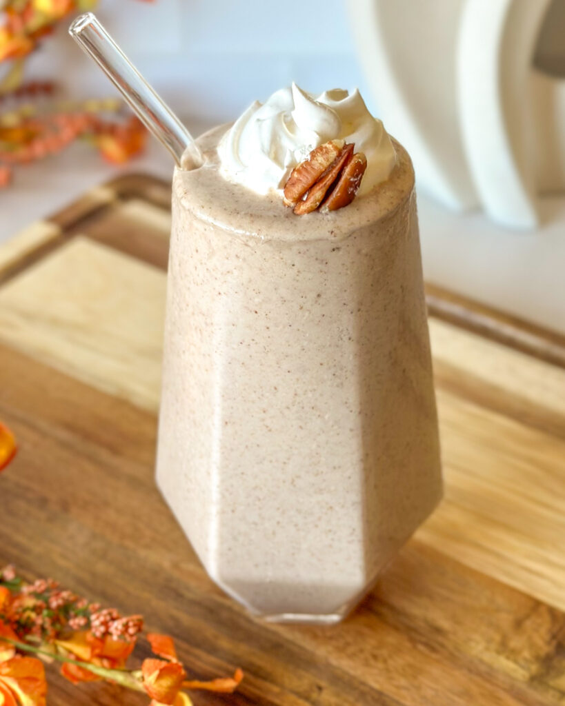 Finished pecan pie smoothie in a geometric glass with whipped cream and a pecan, surrounded by fall flowers on a wooden cutting board