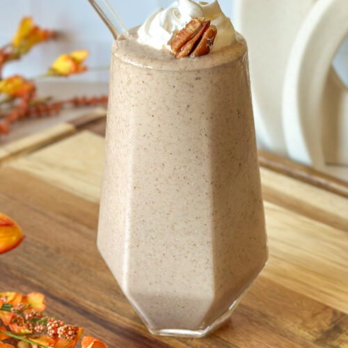 Pecan pie smoothie in a tall geometric glass topped with whipped cream and a pecan, with a glass straw and fall flowers in neutral tones