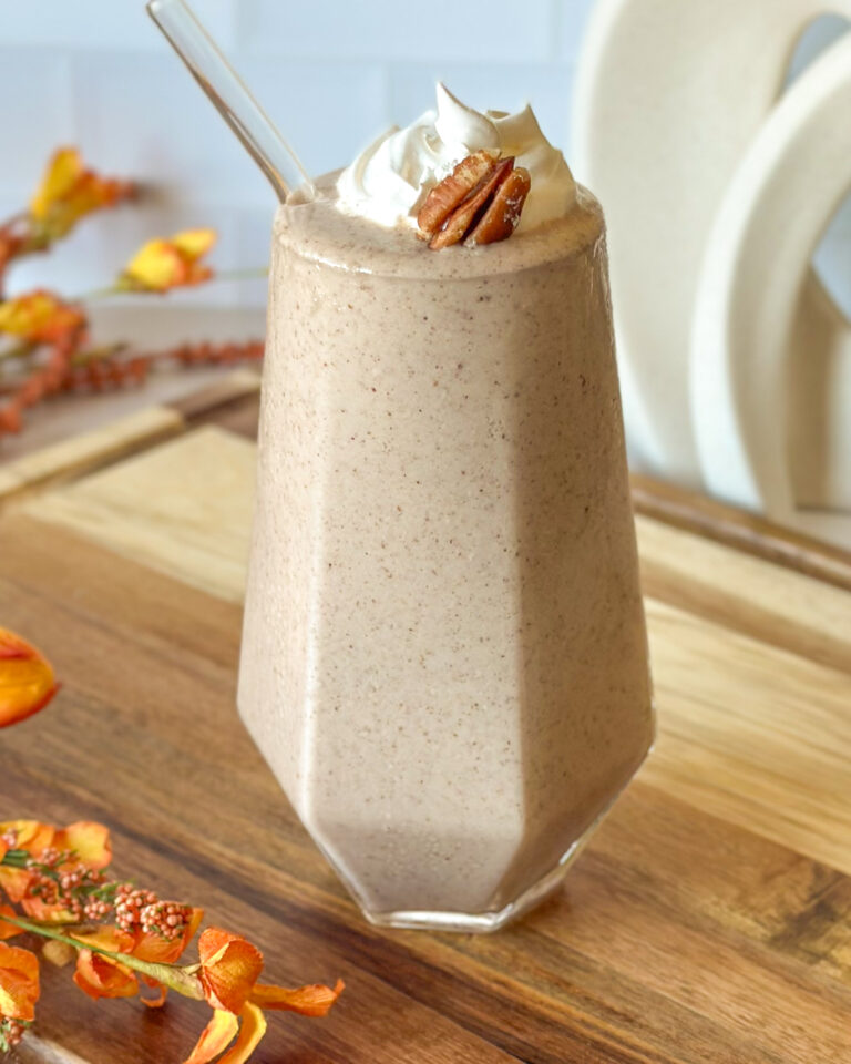 Pecan pie smoothie in a tall geometric glass topped with whipped cream and a pecan, with a glass straw and fall flowers in neutral tones