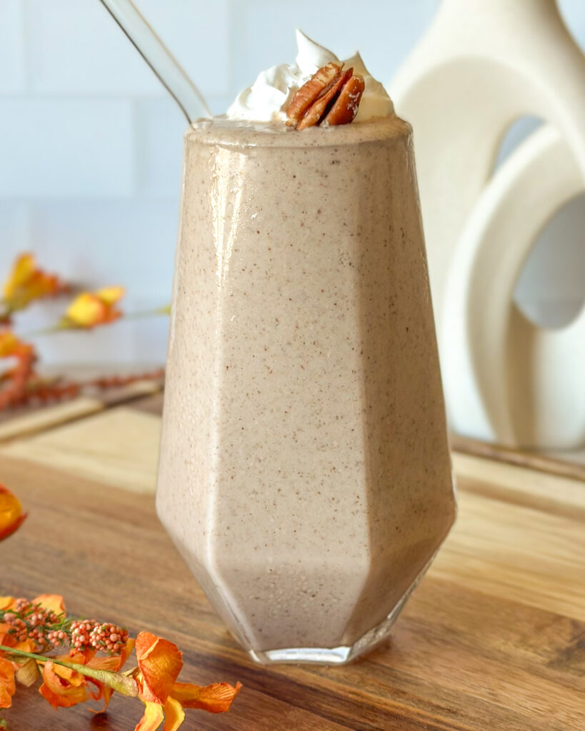 Front-facing pecan pie smoothie in a geometric glass with fall flowers and scattered pecans in the background