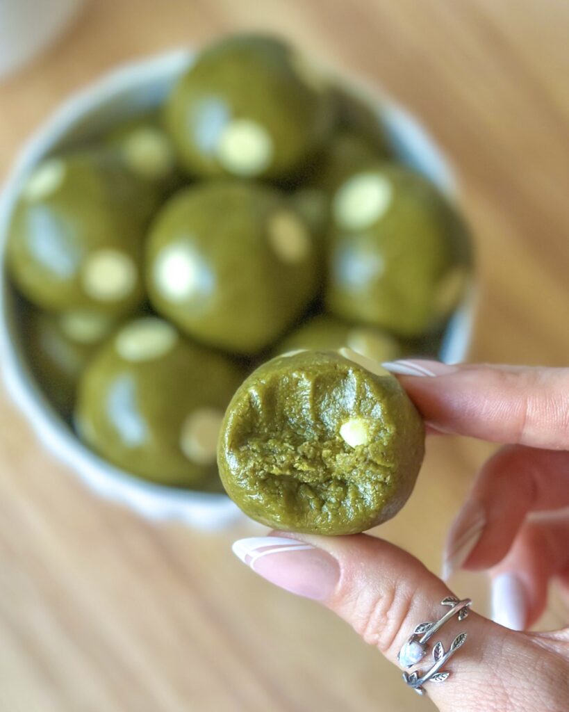 Hands holding a bitten vegan matcha cookie dough ball with a bowl of cookie dough balls in the background