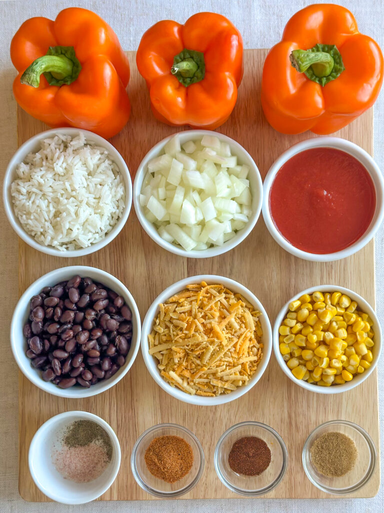 Ingredients for jack-o’-lantern stuffed peppers arranged on a countertop, including orange bell peppers, cooked rice, black beans, corn, onion, garlic, tomato sauce, shredded cheese, olive oil and spices.