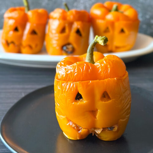 Four orange jack-o’-lantern stuffed peppers filled with cheesy rice, black beans and corn, with their carved Halloween faces visible and one pepper in the foreground on a black plate.