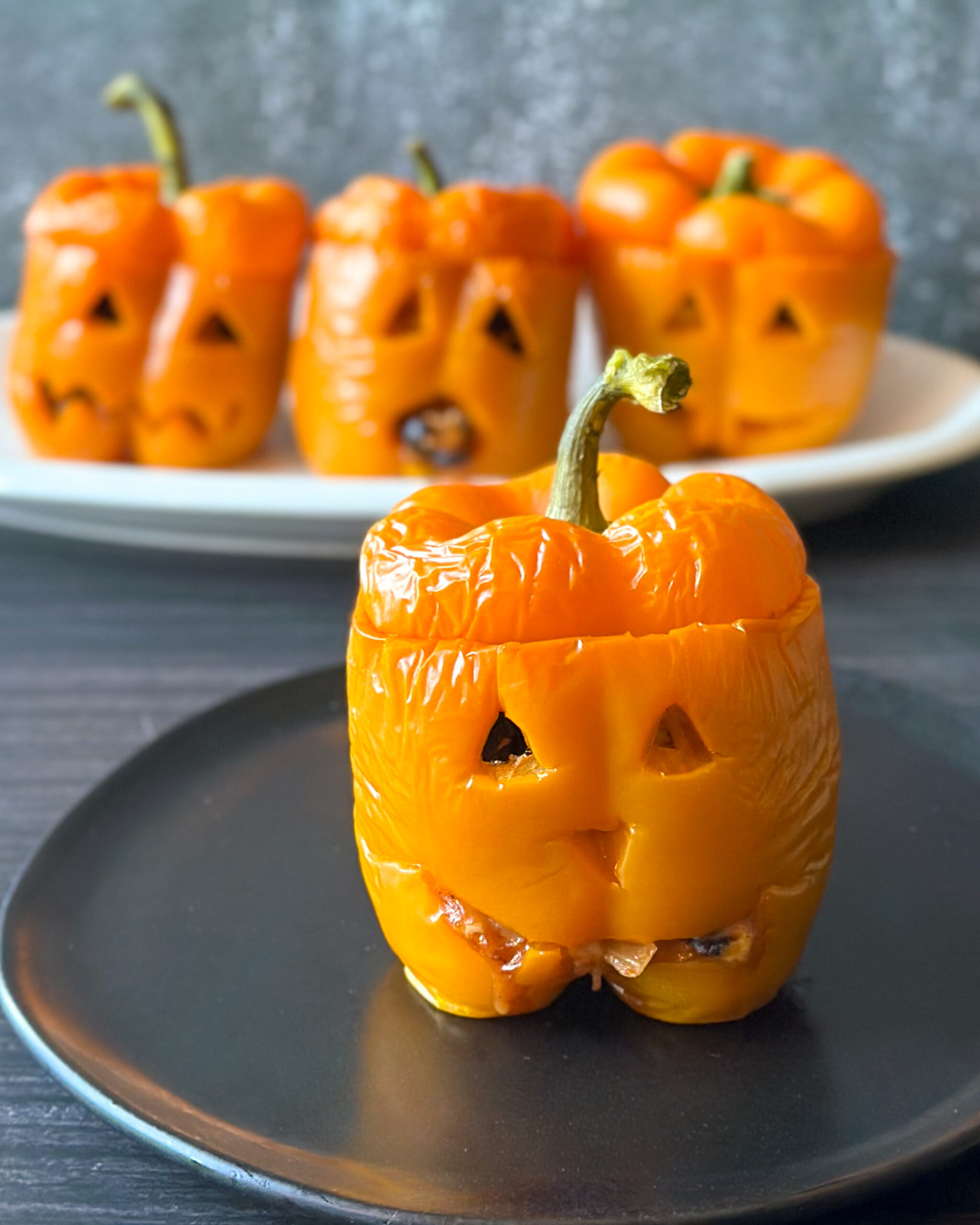 Four orange jack-o’-lantern stuffed peppers filled with cheesy rice, black beans and corn, with their carved Halloween faces visible and one pepper in the foreground on a black plate.