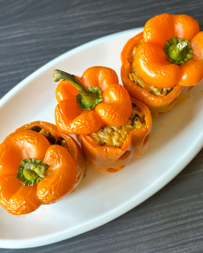 Overhead view of four glossy orange jack-o’-lantern stuffed peppers arranged on a white platter, showcasing their golden tops and cheesy filling.