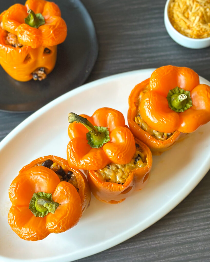 Overhead view of four cheesy jack-o’-lantern stuffed peppers on a white plate, with one pepper’s funny face showing filling spilling through its round mouth and a bowl of shredded cheese nearby.