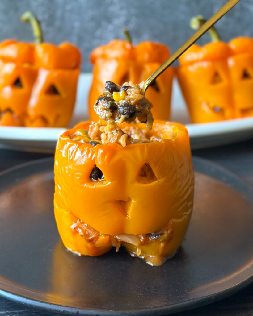 Spoonful of cheesy rice, black bean and corn filling held above jack-o’-lantern stuffed peppers, showing a carved pumpkin face with triangle eyes, an upside-down triangle nose and a toothy grin.