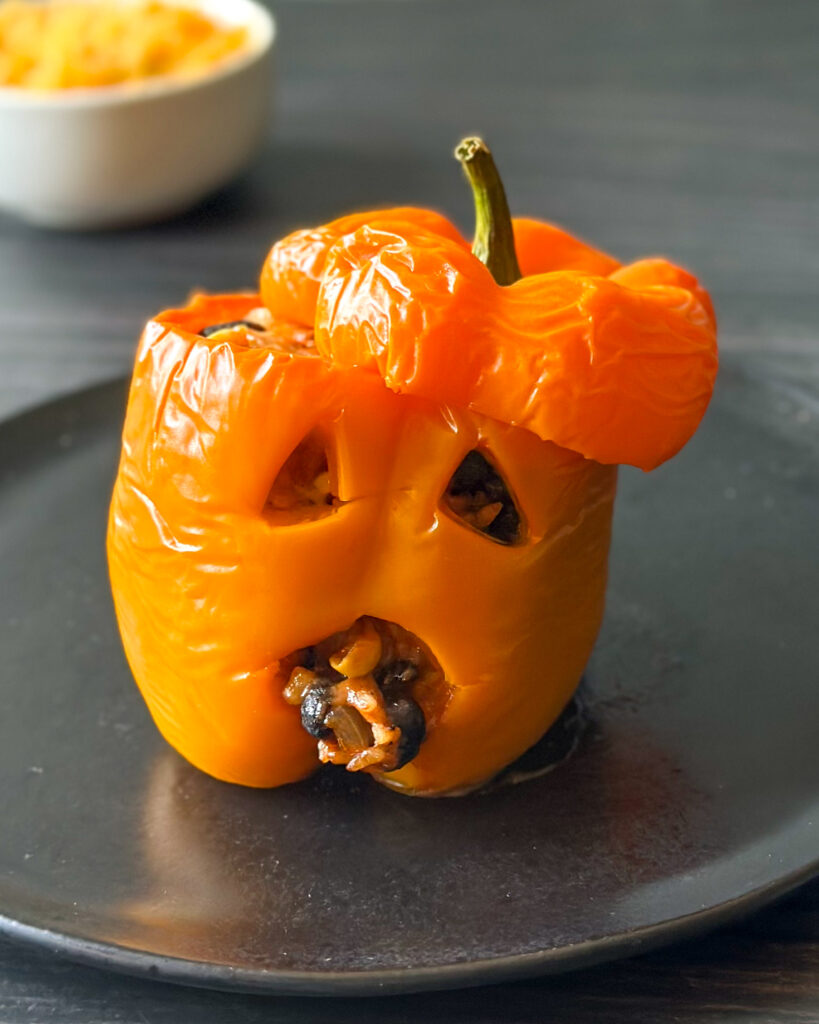Close-up of a cheesy jack-o’-lantern stuffed pepper with triangle eyes and a round mouth filled with rice, black beans and corn, served on a black slate plate with shredded cheese in the background.